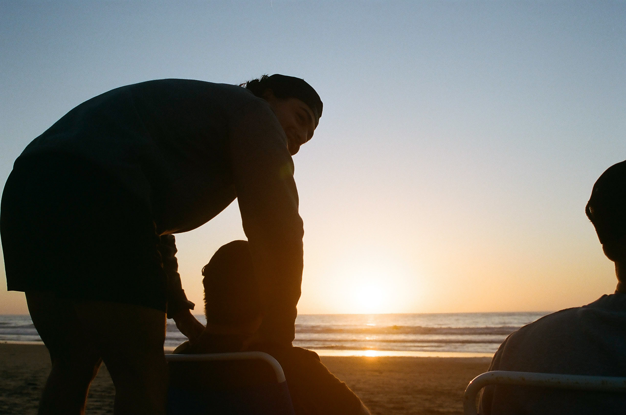 Two friends pictured on the beach in San Diego: one sitting in a beach chair in the sand watching the sky and waiting for the sunset, the other standing over him from behind.