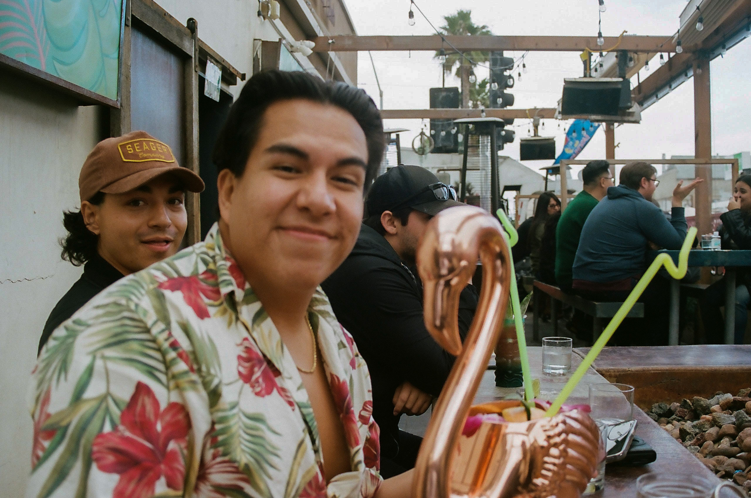 One friend smiling for the picture along with his specialty drink served in a large, copper, flamingo-shaped cup; another friend seated next to him seen photobombing.