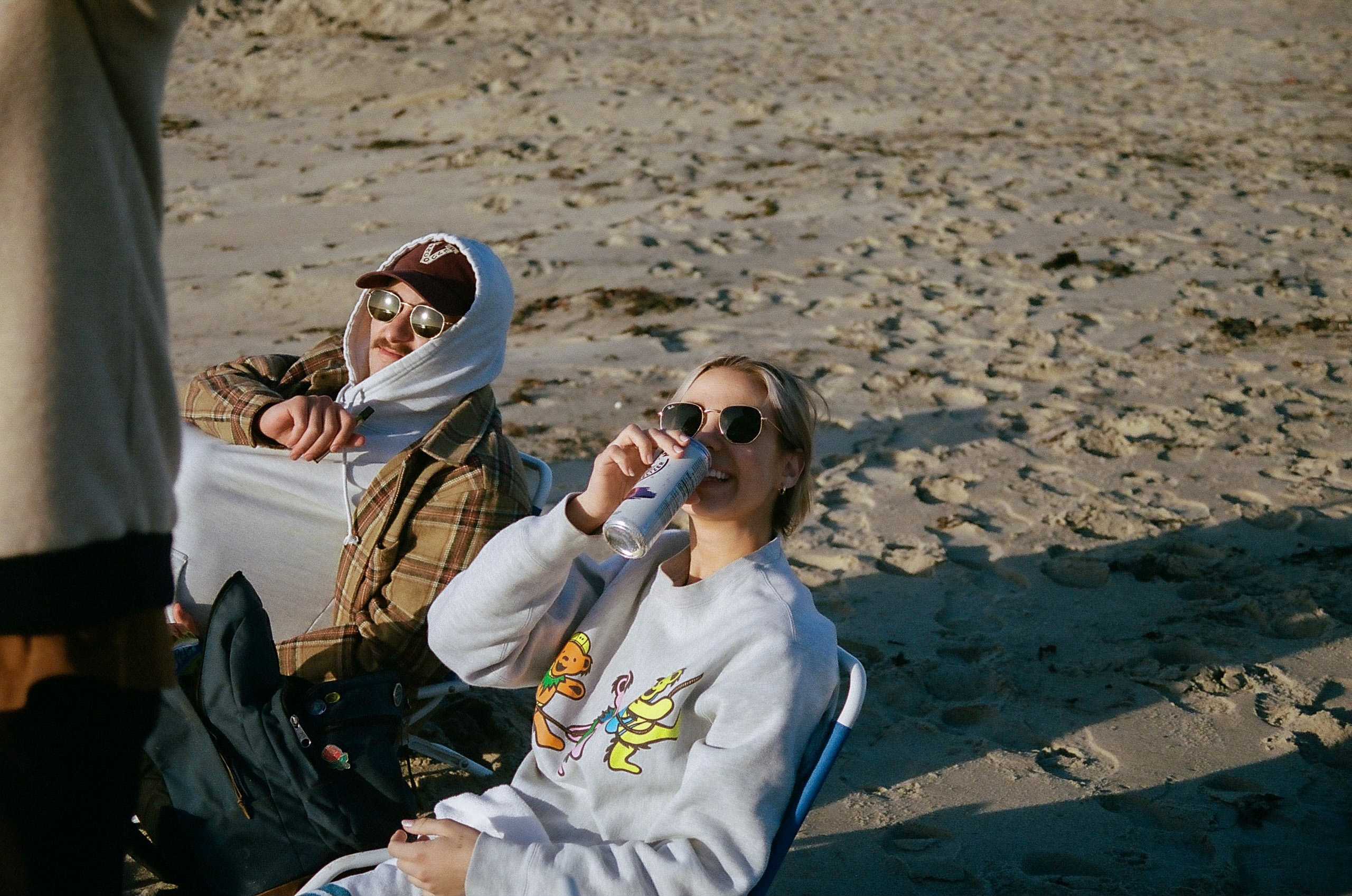 Two friends sitting on beach chairs in the sand; one smiles with her drink in hand, the other bundles up under his jacket and hoodie to stay warm.