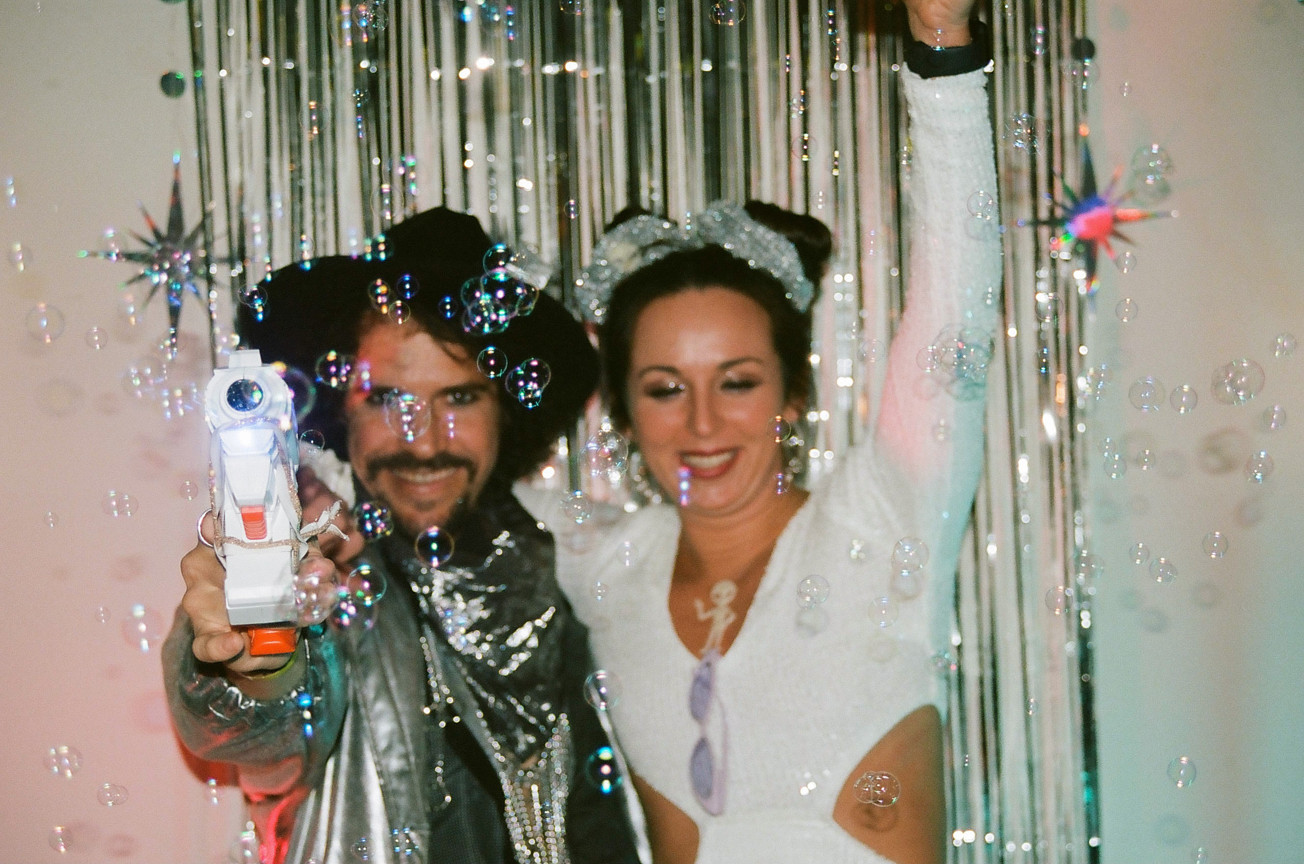 Two partygoers smile and pose in front of decorations; one uses buble gun toy to blow bubbles.