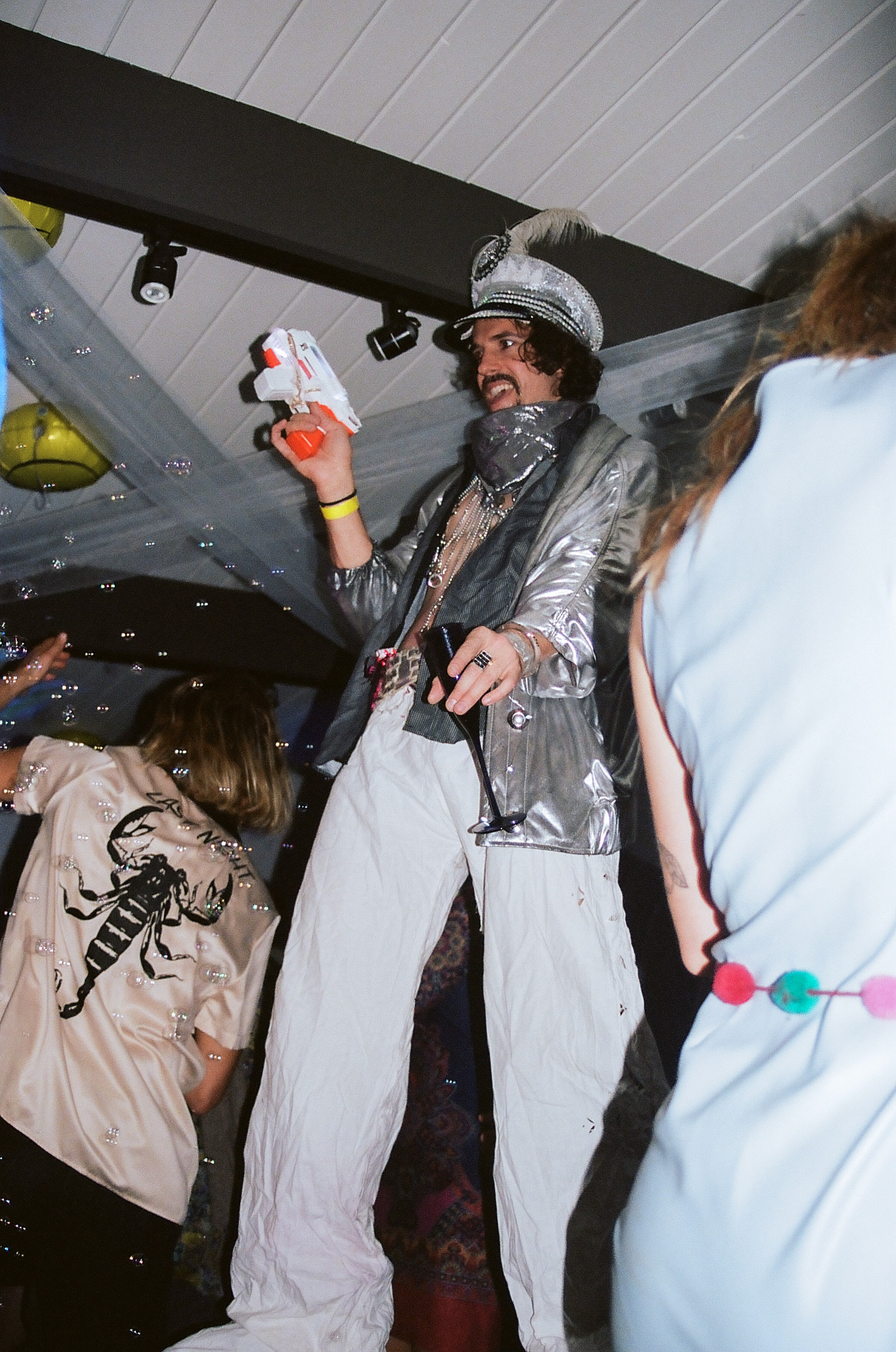 Man on walks through dancefloor on stilts with a blowing bubbles with a bubble gun toy in hand.