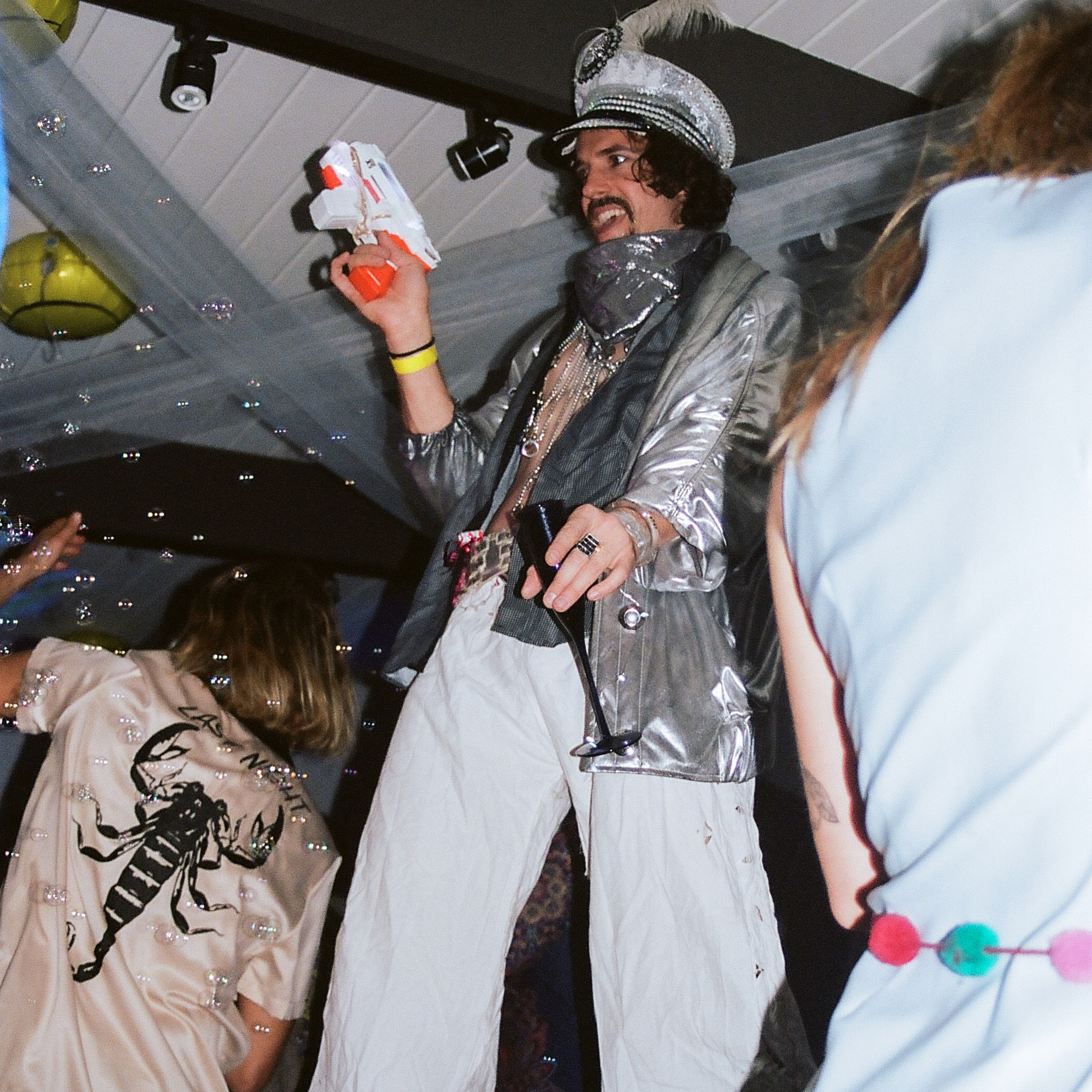 Man walks through the party dancefloor on stilts, champange flute and bubble gun in hand.