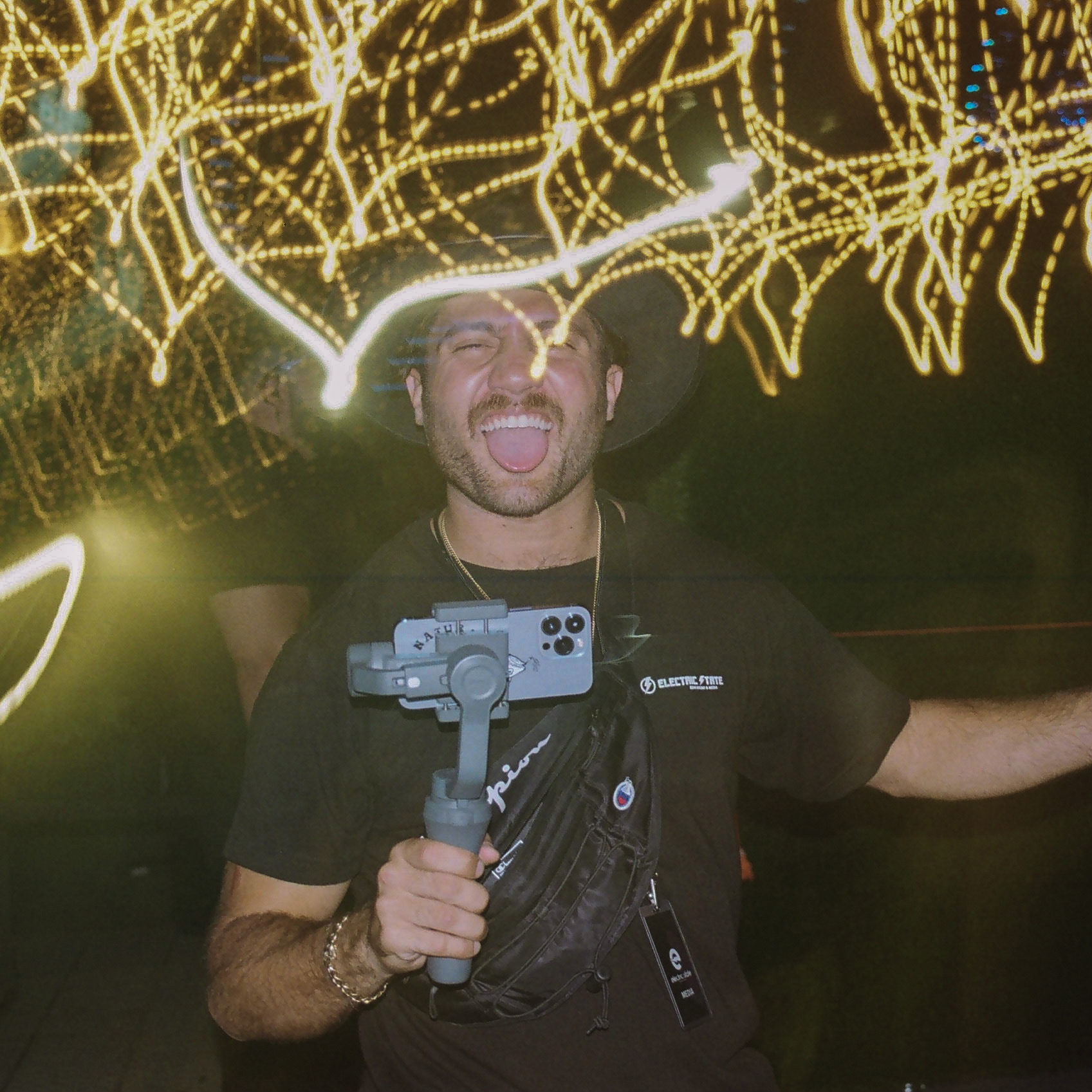 Event videographer poses for a picture; camera's exposure settings creates motion blur of surrounding lights around him.